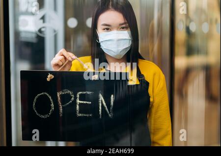 Portrait of a happy waitress standing at restaurant or cafe entrance wearing medical mask on her face. Young Asian woman showing open sign in her small business shop and invites clients Stock Photo