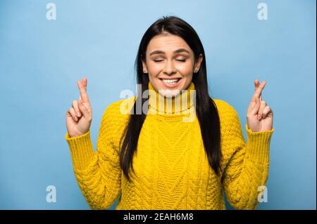 Photo of anxious caucasian girl feel nervous about her dream work, exam, or vacation, cross fingers bite lips and closed eyes, stands on isolated blue background. Stock Photo