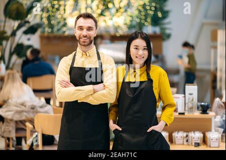Teamwork. Portrait of two friendly waiters. Attractive caucasian guy and beautiful asian girl in black aprons stand inside a restaurant, cafe or bar and smiling Stock Photo
