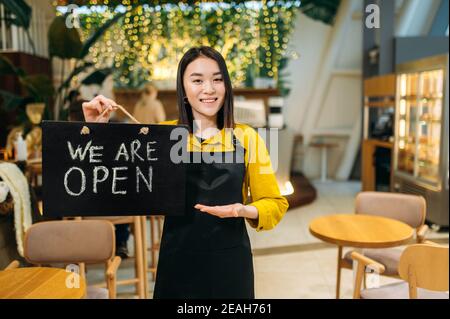 We are open. Portrait of a happy pretty Asian waitress standing indoors of a restaurant or cafe wearing black apron holds OPEN signboard, and friendly smiling Stock Photo