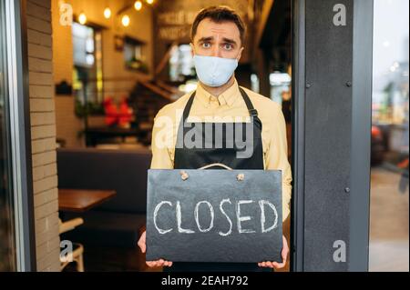 Sad waiter, barista or small business owner. Unhappy man in a medical protective mask and a black work apron stands at the entrance to a cafe, restaurant or bar and holds a CLOSED sign Stock Photo