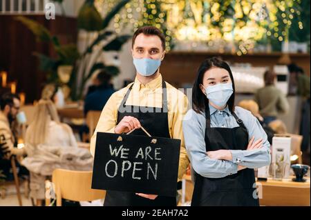 Welcome. Two friendly waiters wearing protective masks. Satisfied young man and asian young woman in black aprons stand inside a restaurant, cafe or bar and showing signboard OPEN Stock Photo
