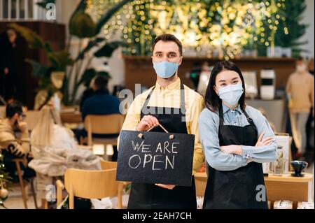 We are OPEN. Two friendly waiters wearing protective masks. Attractive caucasian guy and beautiful asian girl in black aprons stand inside a restaurant, cafe or bar and showing sign OPEN Stock Photo
