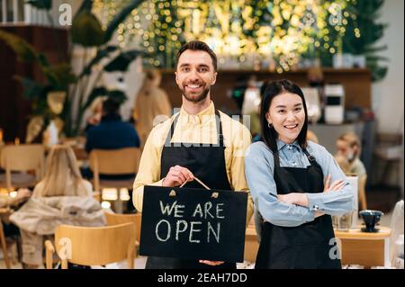 We're OPEN. Two waiters standing indoors. Attractive bearded man and cute asian young woman wearing working black aprons stand inside a restaurant, cafe or bar, showing signboard OPEN, with friendly Stock Photo
