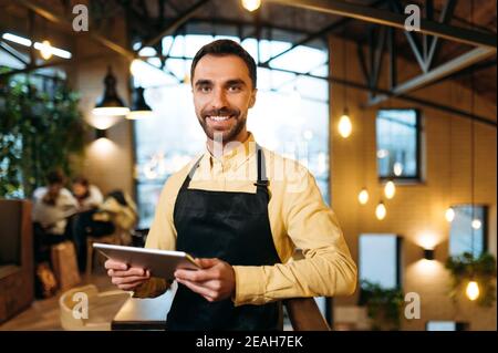 Attractive happy caucasian male waiter or barista wearing black apron stands inside a restaurant, cafe or bar, holds a menu and with friendly smile looks at the camera Stock Photo