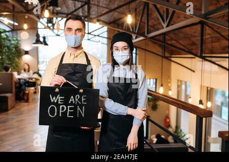 We are OPEN. Two friendly multiracial waiters wearing protective masks. Attractive caucasian guy and beautiful asian girl in black aprons stand inside a restaurant, cafe or bar and showing signboard Stock Photo