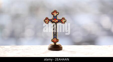 A stark image of a detailed colorful crucifix ornament on a lightly patterned satin sheet with a plain heavily blurred bokeh background. Christianity Stock Photo