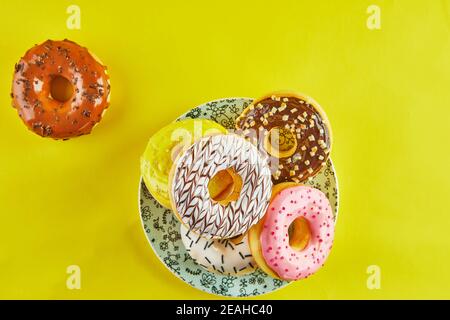 Multicolored donuts with icing and splashes on a blue plate on a yellow background. Flat lay Stock Photo