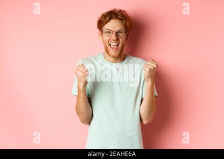 Excited young man winning prize, shouting with joy and triumph, making fist pump and saying yes, standing over pink background Stock Photo