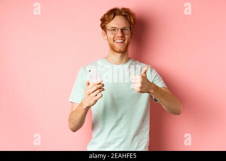 Happy young man with red messy hair, showing thumbs-up while using mobile phone, smiling and praising application, standing over pink background Stock Photo