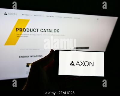 Person holding mobile phone with logo of American weapon manufacturer Axon Enterprise Inc. on screen in front of webpage. Focus on cellphone display. Stock Photo