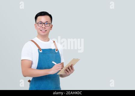 Young Asian man startup business owner wearing apron taking order on clipboard and looking at camera isolated on light gray backrground Stock Photo