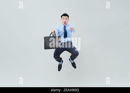 Shocked excited young Asian businessman with briefcase jumping in mid -air and pointing finger at camera isolated on light gray background Stock Photo