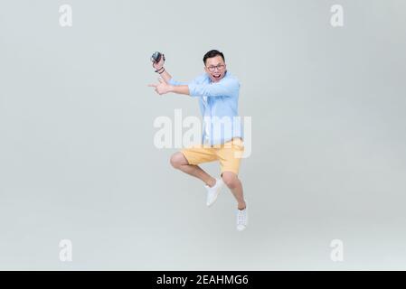 Young Asian man with camera jumping and pointing hand to empty space on light gray backgground Stock Photo