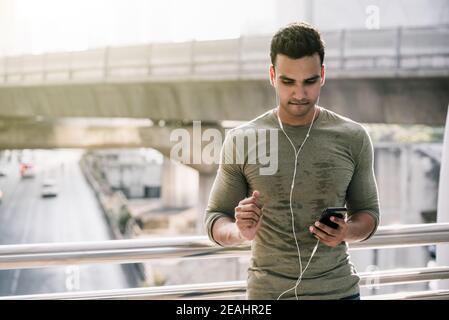 Young handsome Indian man in sweaty top resting after jogging outdoors and listening to streaming music on smartphone against urban traffic background