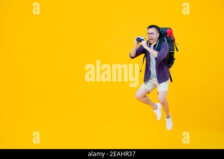 Young handsome happy Asian man in tourist clothes with backpack taking photo and smiling while jumping beside copy space isolated on yellow background Stock Photo