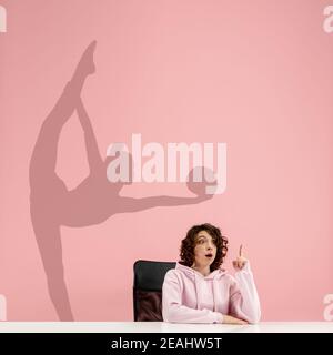 Young caucasian woman dreaming about future in big sport during her work in office. Shadow, silhouette of female rhythmic gymnast on the wall. Becoming a legend. Inspiration, aspiration. Copyspace. Stock Photo