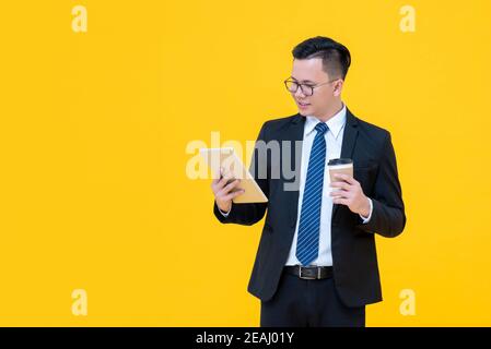 Focused Asian businessman looking at tablet computer while drinking coffee isolated on yellow background Stock Photo