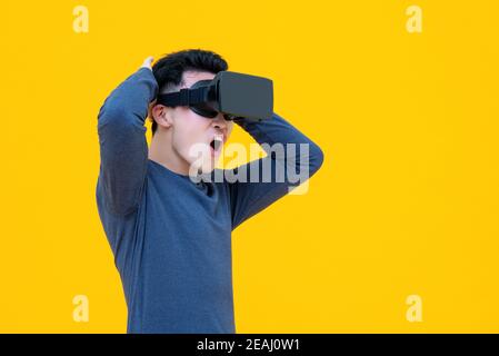 Excited Asian man enjoying watching  3D simulation video from virtual reality or VR glasses on yellow background Stock Photo