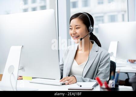 Smiling beautiful Asian woman wearing microphone headset working in call center office as a telemarketer Stock Photo
