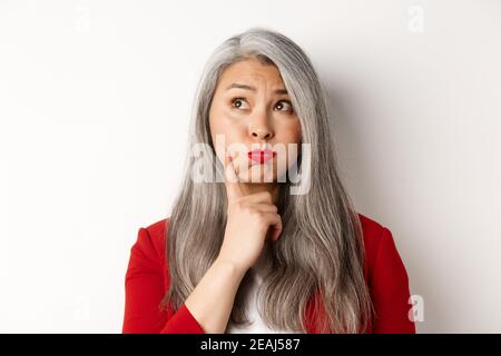 Close up of indecisive asian senior woman making choice, pouting and looking thoughtful at upper left corner, standing against white background Stock Photo