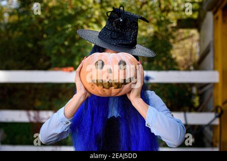 A girl dressed as a witch covered her face with a pumpkin with an evil face drawn in the style of a Halloween celebration Stock Photo