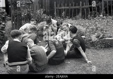 1950s, historical, 'hanging out with friends, a group of boys, some  teenagers, others slightly younger, sitting in a suburban back garden having a chat and a laugh together, England, UK. All are wearing short trousers and some have cotting tank tops over their shirts. Tank tops are a sleeveless jumper and knitted ones made of ribbed cotton were a common attire for males and young men in particular in this era. Stock Photo