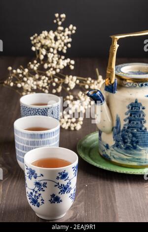 View of three blue cups with tea, white flowers and china teapot, selective focus, on wooden table, vertical Stock Photo