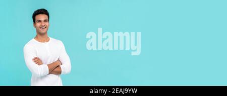 Smiling happy Indian man with arms crossed in plain white t-shirt isolated on light blue banner background with copy space Stock Photo