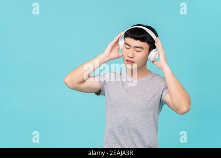 Asian man in casual gray t-shirt wearing headphones listening to emotional music with eyes close isolated on light blue background Stock Photo