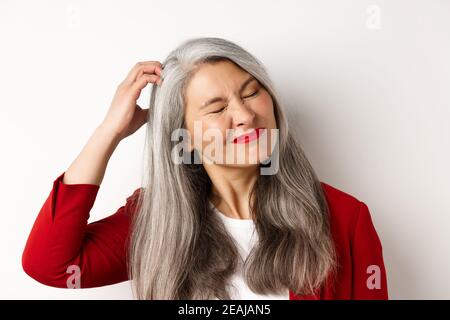 Close up of senior asian woman scratching head and looking bothered by itching, standing over white background Stock Photo