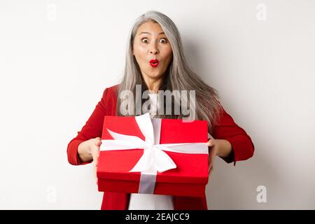 Surprised asian senior woman receiving present on mother day, holding red box with gift and looking amazed at camera, white background Stock Photo