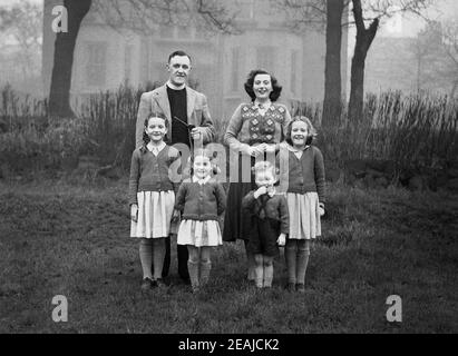 1950s, historical, a family photo, standing outside on grass in the church grounds, a vicar with his wife and four young children with little girls in the school uniform., England, UK. The vicar, wearing his clerical collar is holding a long pipe. Stock Photo