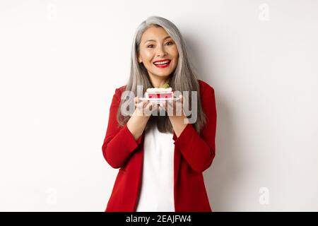Beautiful asian grandmother holding piece of cake and smiling, standing in red blazer over white background Stock Photo