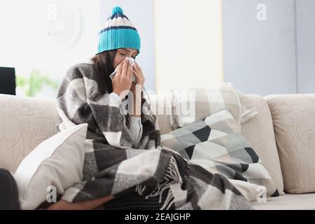 chilled woman on sofa in blanket and hat holds handkerchief Stock Photo
