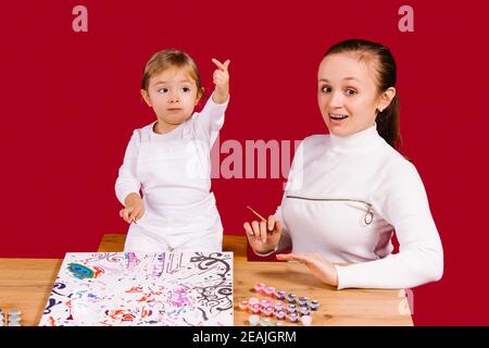 Caucasian blond woman looking at the camera with her daughter on a red background during her favorite pastime. Painting by numbers and bright colors.