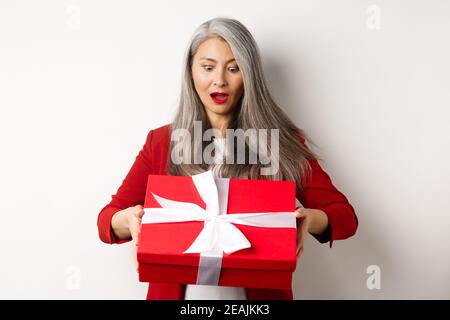 Surprised asian senior woman receiving present on mother day, holding red box with gift and looking amazed, white background Stock Photo