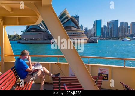 A man wearing a Covid face mask sits and reads a newspaper in the late afternoon sun on the stern deck of a Manly Ferry as it passes the Opera House Stock Photo