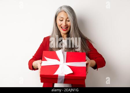 Happy elderly woman with grey hair, receive present, looking at red gift box and smiling surprised, standing over white background Stock Photo