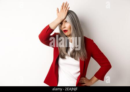Annoyed asian office lady in red blazer roll eyes and facepalm, standing bothered and irritated over white background Stock Photo