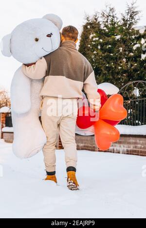 Young man from behind hugs holding big teddy bear soft toy gift heart air balloons on this special day surprising girlfriend. romantic surprise for Bi Stock Photo