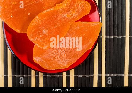 Large slices of dried mango in a red saucer on a bamboo mat, close up. Stock Photo