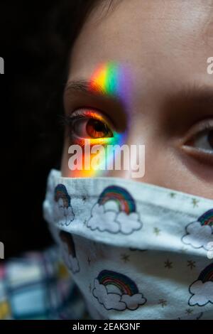 Closeup of little girl with colorful mask and rainbow colored sunlight on her eye, looking to the camera; concept: pandemic, Covid19, hope Stock Photo