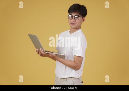 happy cheerful young asian man nerd student dressed casually wearing eyeglasses holding laptop computer isolated on yellow studio background Stock Photo