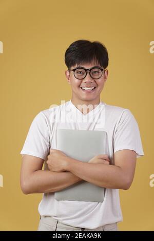 happy cheerful young asian man nerd student dressed casually wearing eyeglasses holding laptop computer isolated on yellow studio background Stock Photo