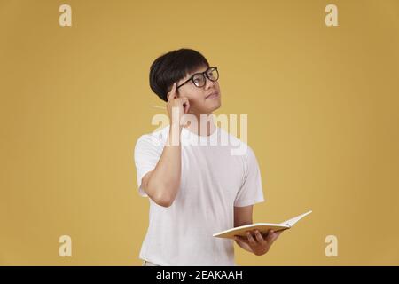 thoughtful young asian man student dressed casually wearing eyeglasses holding notebook thinking isolated on yellow studio background Stock Photo