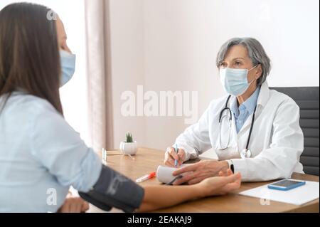 Senior mature female doctor wearing a lab coat and face mask, using sphygmomanometer with a stethoscope, checking the results of blood pressure test Stock Photo