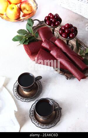 Cherry pastille is rolled into a cone. Two cups of coffee in vintage dishes. Vertical photo. Cherries and apples are in a bowl of Nickel silver. Stock Photo
