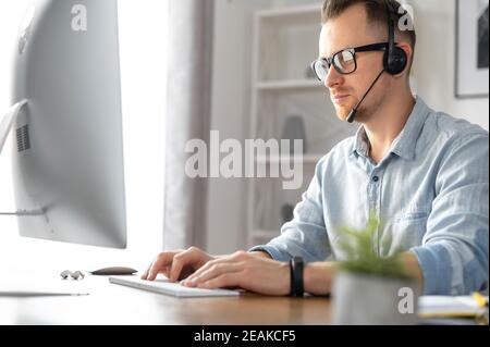 Side view of young male wearing formal shirt using a headset and contemporary PC in the office, intelligent guy talking online and typing sitting at Stock Photo