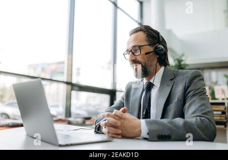 Mature male manager in headset and formal suit sits at the desk, work as call center operator, communicate with clients. Caucasian businessman is consulting people, support service concept Stock Photo